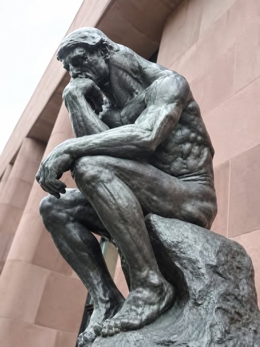 A low-angle photograph of a bronze 'Thinker' statue sitting on a rock, set against a stone building facade.