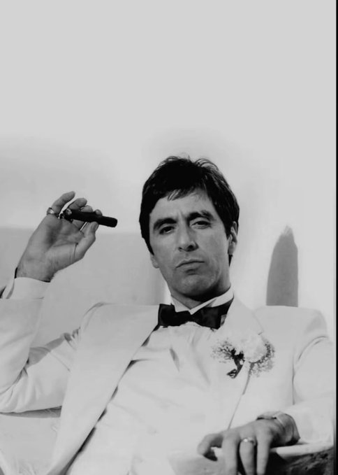 A black and white portrait of a man in a white tuxedo jacket with a black bow tie and boutonnière, holding a cigar near his face while reclining against a light-colored background.