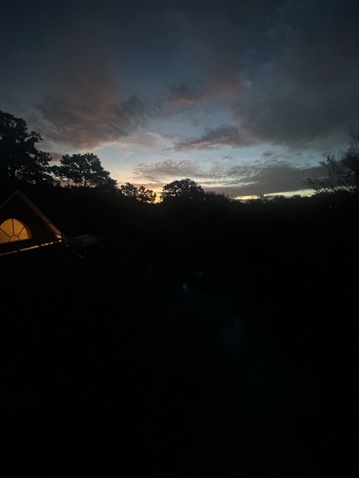 A serene twilight landscape featuring a dark silhouette of trees and a small illuminated tent against a sky streaked with pink, blue, and orange sunset hues.