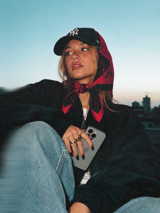 A photograph of a young woman wearing a black NY Yankees cap and a red and black patterned scarf, holding an iPhone with a city skyline at dusk in the background.