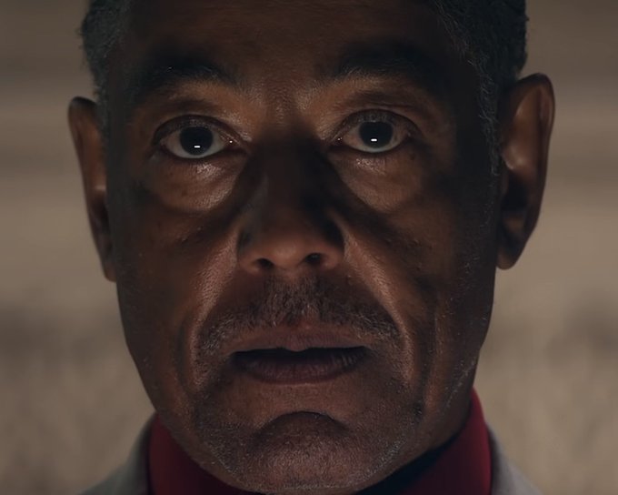 Close-up portrait of an older man with a serious expression, short graying hair, and stubble, wearing a red collared shirt, with dramatic lighting on his face.