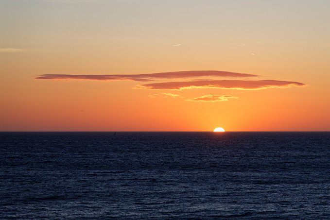 A scenic view of a sunset over a calm ocean, with the sun dipping just below the horizon and a long cloud formation stretched across the sky.