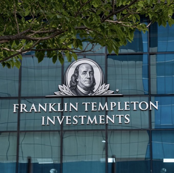 A photograph of a glass building exterior featuring the Franklin Templeton Investments logo, which includes a portrait of Benjamin Franklin encircled by laurels, with green tree leaves in the foreground.