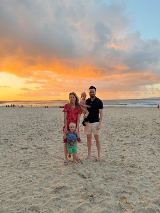 A family of four—two adults and two young children—poses together on a sandy beach at sunset, with vibrant orange and pink clouds stretching across the sky over the ocean, capturing a cherished family moment that the poster values above market gains.