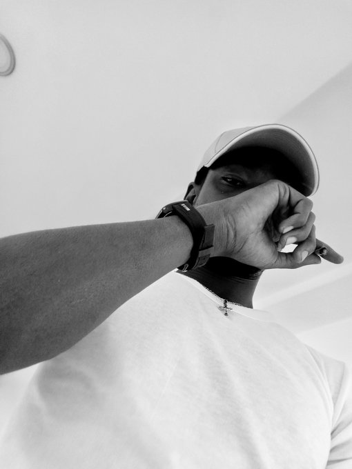 Black and white low-angle photo of a man wearing a white cap and white t-shirt, with his hand near his face, wearing a watch.