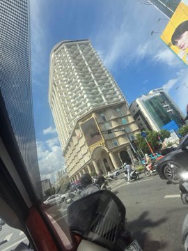 A street-level photo taken from a vehicle showing a tall multi-story building with distinctive architecture in an urban Asian city setting, with motorcycles and pedestrians visible at street level under a partly cloudy sky.