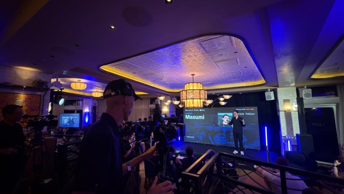 A dimly lit event venue with purple/blue lighting where a presenter named 'Masumi' is on a screen being filmed by audience members.