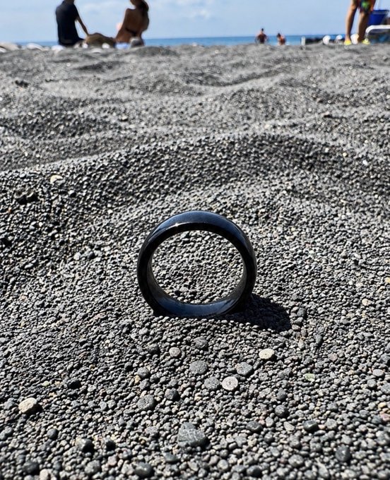 Close-up photo of a black band/ring standing upright on dark gray pebbly beach sand, with blurred beachgoers visible in the background.