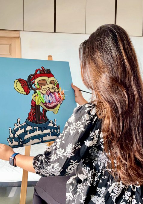 A woman with long brown hair painting a colorful abstract portrait on a blue canvas resting on a wooden easel.