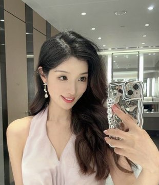 A young woman with long dark hair takes a mirror selfie in a modern interior while wearing dangling earrings and a white halter top, holding a phone with a decorative case. The glamorous, stylish self-portrait pairs with the "$JACKET" meme post, presenting the transformation—taking off one outfit and donning another—as a fashionable, confident reveal.