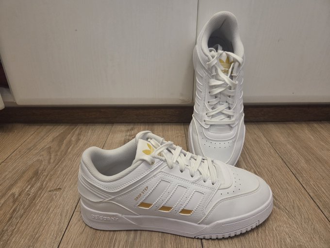 A pair of white Adidas sneakers with gold accents, including the Adidas trefoil logo and three gold stripes, displayed on a wooden floor against a white wall.