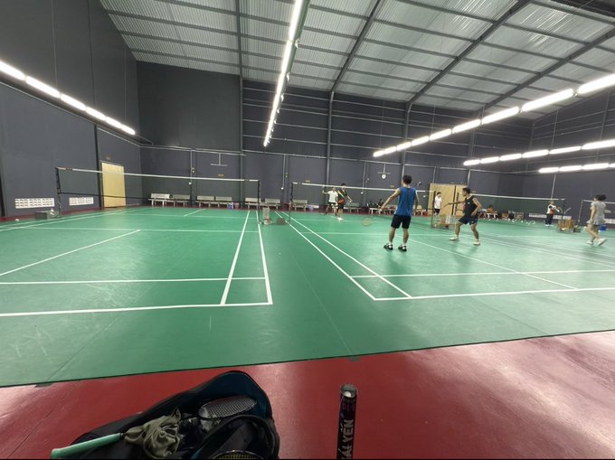 An indoor badminton court with green flooring where several people are playing, with a badminton racket visible in the foreground.