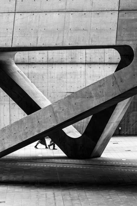 A black and white photograph captures a striking modern concrete sculpture with intertwining curved forms, set against a perforated wall, with two small figures walking beneath its massive archway in Seoul. The bold geometric lines and dramatic shadows complement the "GM Friends Seoul, 2023" theme, emphasizing contemporary architecture and human scale within an urban landscape.