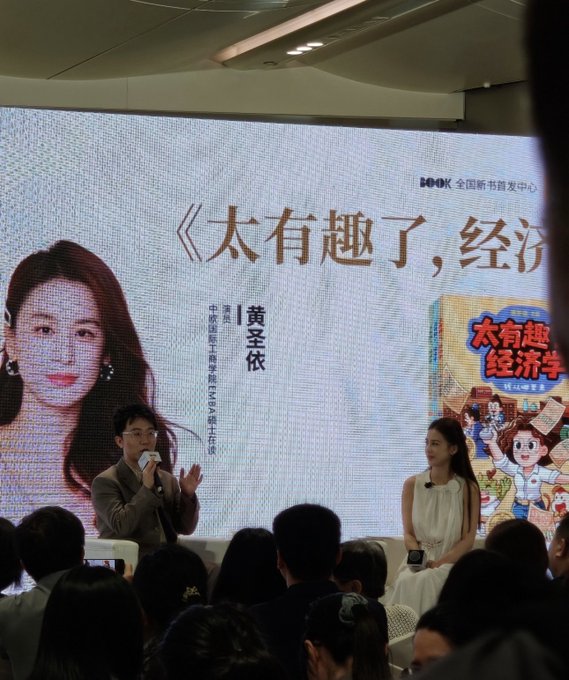 An indoor book event where a man with a microphone speaks next to a seated woman, in front of a large screen displaying a book cover and Chinese text '太有趣了, 经济' by author 黄圣依, with 'BOOK' visible in the corner.
