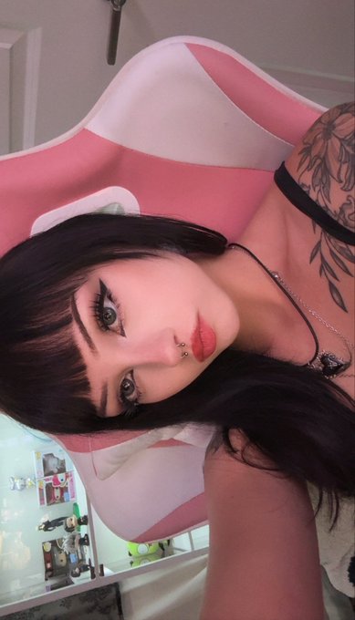 A young woman with a septum piercing and floral shoulder tattoo poses for a selfie in front of a pink gaming chair, with a "gm freaks" greeting posted to match her casual, aesthetic vibe.