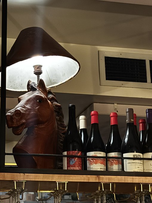 A bronze horse head lamp sits beside a row of wine bottles, including a Chambolle Musigny, on a shelf. The image ties to the caption's message of doubling down on conviction—symbolizing the rider charging forward with renewed determination.
