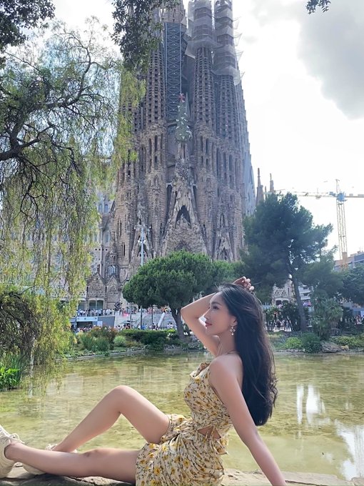 A young woman in a yellow floral dress posing in front of the Sagrada Família basilica in Barcelona, with a pond and trees in the foreground.
