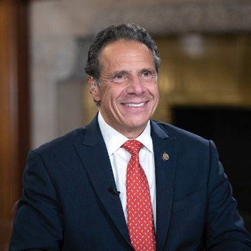 The image shows Andrew Cuomo, former New York Governor, smiling in a dark suit with a red patterned tie and a lapel pin during what appears to be a televised interview. The photo relates to the announcement of his appointment to OKX's board and as co-chair of the crypto exchange's joint venture with NYSE parent ICE, marking his continued influence in business and finance after his governorship.