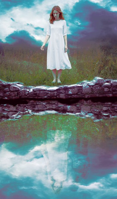 A woman with red hair wearing a flowing white dress stands on a stone wall in a grassy field, with her surreal reflection appearing in vibrant turquoise and purple water below.
