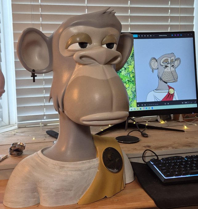 The image features a large, gray monkey statue sitting next to a computer monitor with an animated character drawing or painting displayed on its screen. The monkey's head is tilted upwards, giving it a curious expression as if observing something interesting on the monitor. The scene suggests that someone might be working on a project related to the animated character or simply enjoying some creative inspiration from the monkey statue.