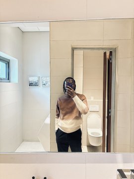A mirror selfie of a person wearing a brown and white sweater and glasses, taken in a bathroom with a urinal visible in the reflection.