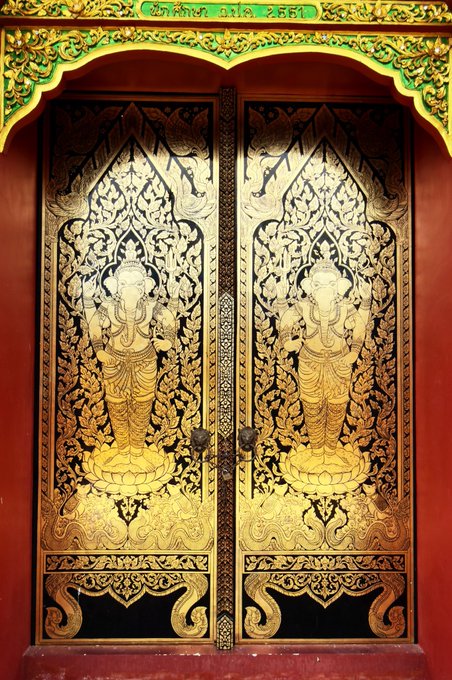 An ornate pair of red temple doors adorned with intricate gold leaf designs depicting deity figures and traditional Thai patterns within a decorative archway.