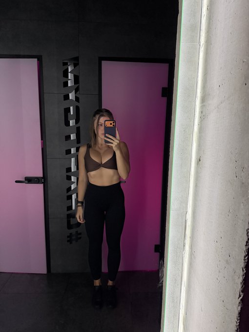 A mirror selfie shows a woman wearing a brown sports bra and black leggings, standing in front of pink doors with 'PYHCKD #FRIGHT' painted on the wall.