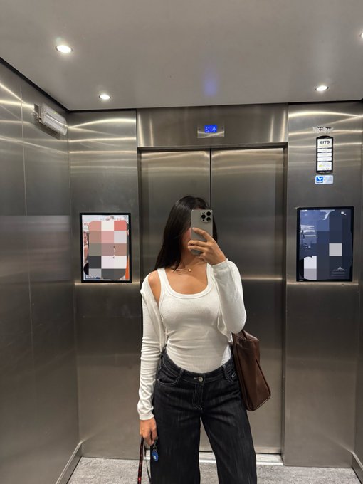 Mirror selfie of a woman in a white top and dark jeans standing in a stainless steel elevator, holding a phone and a brown handbag.