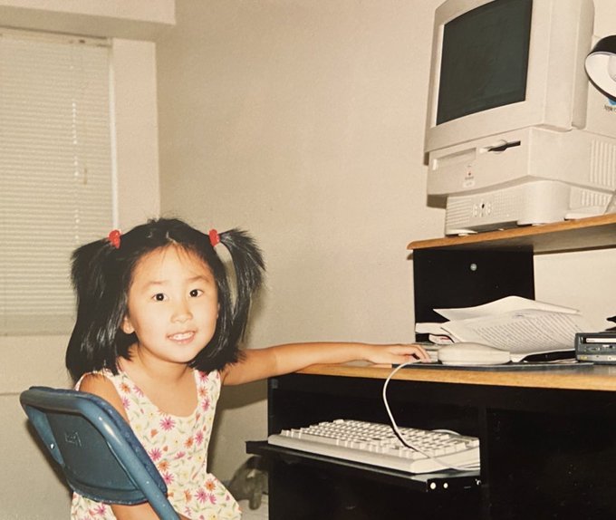 A young Asian girl with pigtails sits at a desk, reaching toward an Apple/Macintosh computer setup from the 1990s, smiling at the camera. The nostalgic image contrasts the innocence of childhood with the post's message about token fraud, suggesting the speaker is defending their real identity or roots against tokenism.