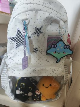 A white backpack decorated with star patterns and featuring a clear front pocket containing plush toys, along with a bat keychain and tags.
