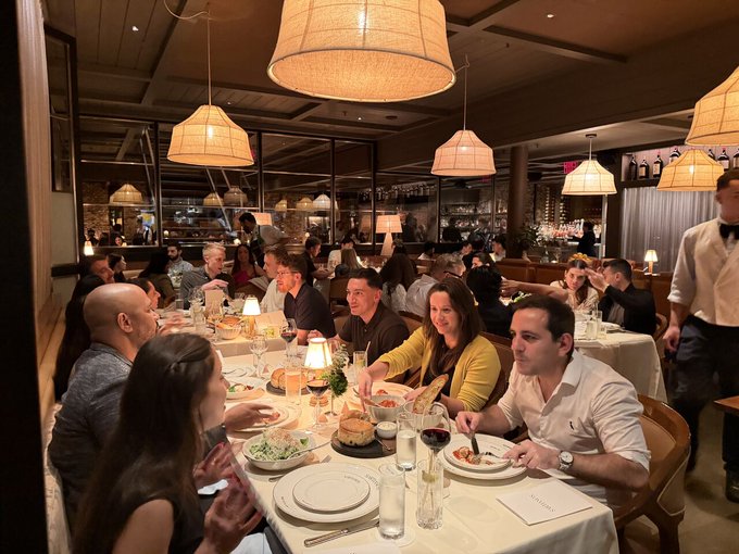 A photo of a lively restaurant scene where a group of people are dining together at a table filled with plates and wine glasses, with a server in a vest and bowtie standing nearby.