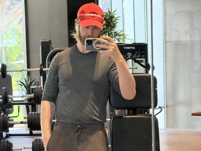The image features a man taking a selfie with his cell phone, wearing a red baseball cap while standing next to some dumbbells at a gym. The caption of the image reads "as a @monad contributor, there is no excuse for being soft ... how will you hold a floor ... how can you pump a bag ... how can you carry a meme this is why all contributors (like you) are shredded gymonad". This message highlights the importance of maintaining physical fitness and encourages gym-goers to stay committed to their workout routines.