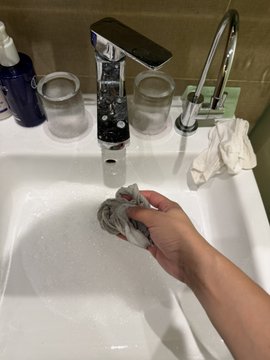 A hand washes a single gray sock in a soapy bathroom sink, with two clear drinking glasses and a soap dispenser nearby, illustrating the travel mishap of having only mismatched socks after a morning run in Singapore.