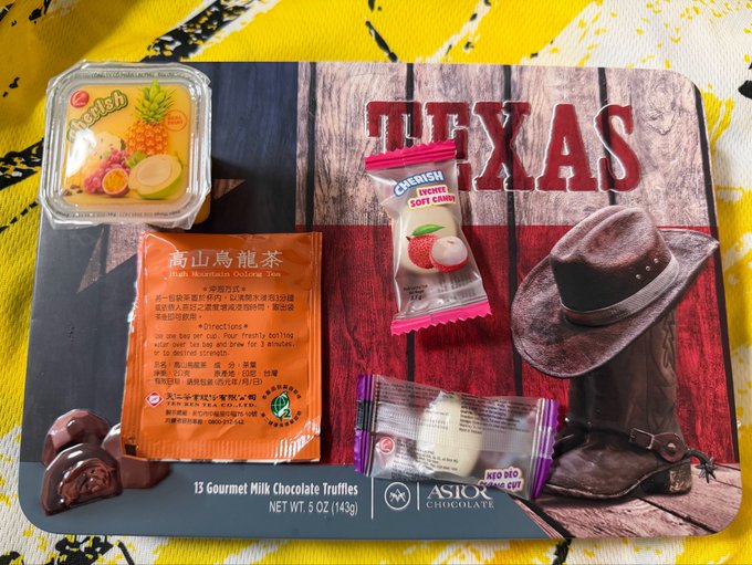 A flat-lay photograph showing various items including a fruit cup, 'High Mountain Oolong Tea' packet, 'Cafresh Lychee Soft Candy', and a 'Texas' themed Astor Chocolate box on a yellow patterned surface.