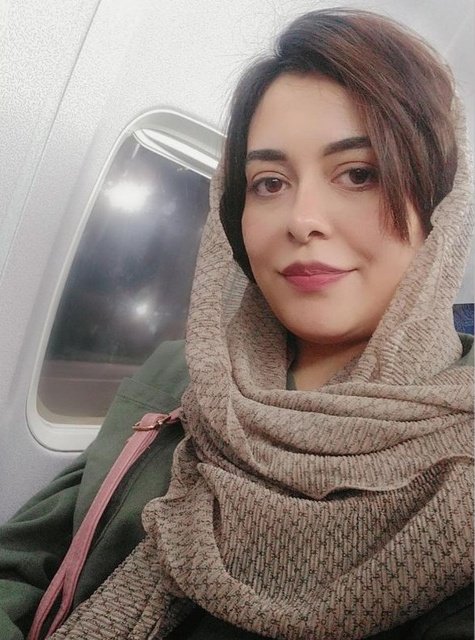 A selfie of a woman with brown hair wearing a beige knit scarf, seated next to an airplane window.