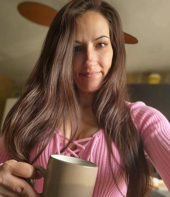 A selfie photo of a woman with long brown hair wearing a pink lace-up sweater, holding a gold/beige ceramic mug in what appears to be a home setting.