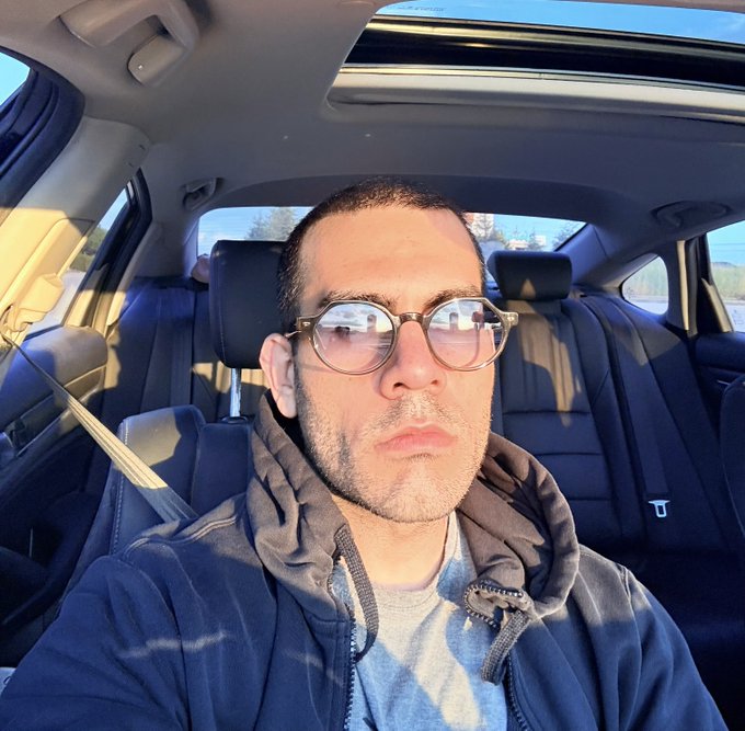 The image features a man wearing glasses, sitting inside a car with his head resting against the window. He appears to be looking out of the vehicle's window or taking a selfie. The photo has been captioned "This guy thinks he can make NFTs great again", which suggests that the man might have an interest in collecting and trading digital artworks, such as NFTs (Non-Fungible Tokens).