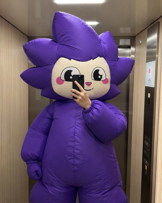 A person inside a large inflatable purple mascot costume takes a mirror selfie with a smartphone inside an elevator.