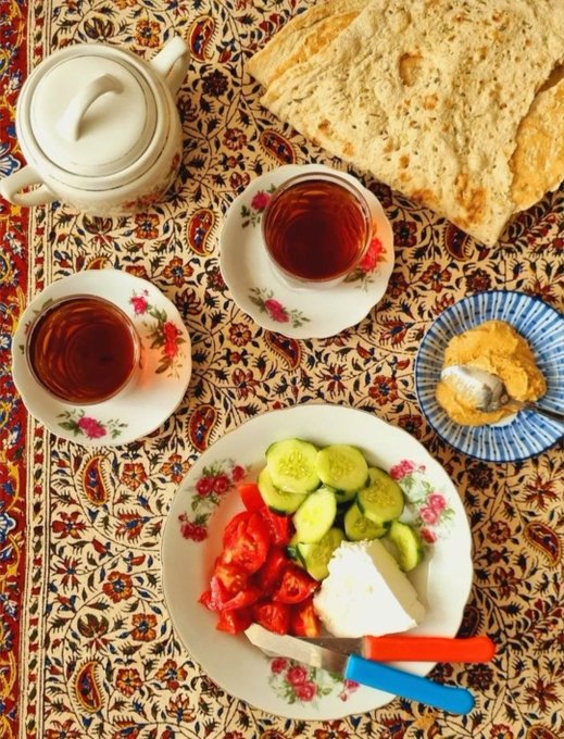 An overhead photo of a Middle Eastern/Mediterranean breakfast spread on a Persian rug, including two glasses of tea in floral saucers, fresh bread, a plate of tomatoes, cucumbers, feta cheese, and a small dish of cream or hummus.
