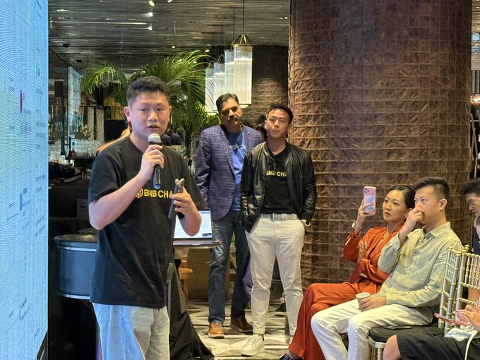 A man in a black 'BIG CHAD' t-shirt speaks into a microphone at an event, with attendees watching, including a man in a 'BIG CHAD' leather jacket and others seated and recording.