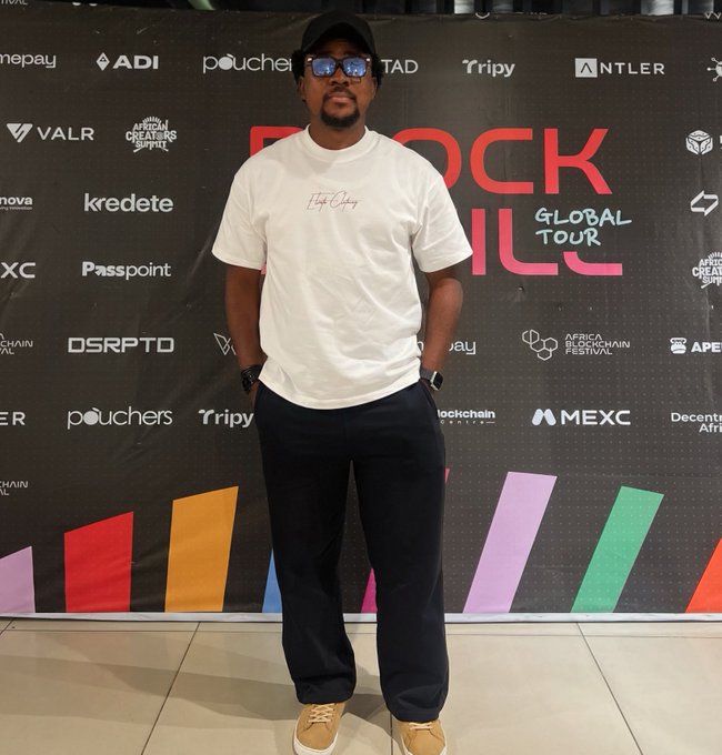 A man wearing a white t-shirt, dark pants, cap, and sunglasses stands in front of a 'BLOCK NIL GLOBAL TOUR' backdrop featuring various sponsor logos.