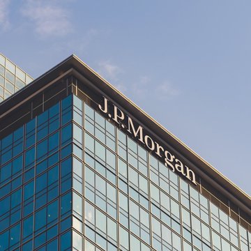 The image shows the upper corner of a modern glass office tower with the "J.P. Morgan" name displayed in large white letters against the blue-tinted facade, set against a clear sky. This serves as a visual identifier for the financial giant referenced in the accompanying post, which discusses JPMorgan's commentary on the CLARITY Act's uncertain legislative prospects.