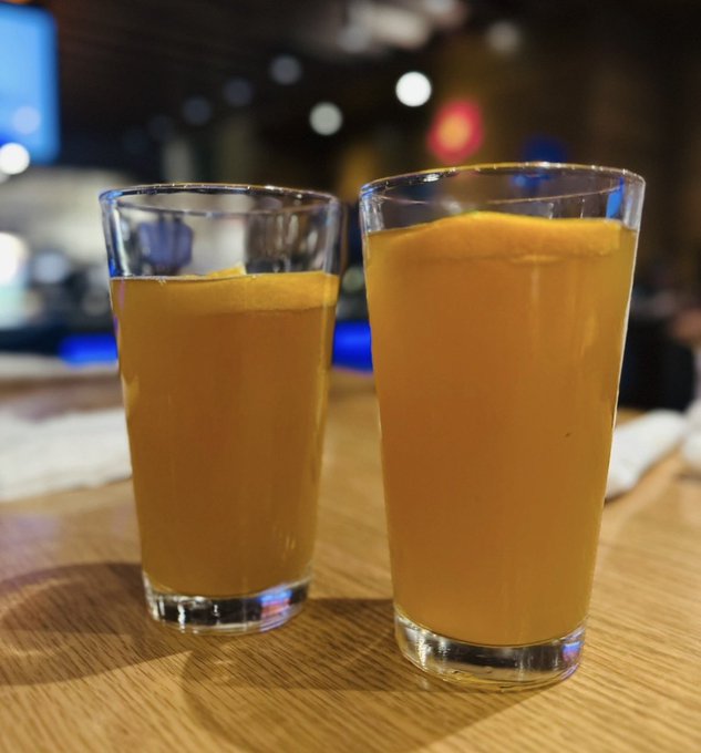Two tall glasses of freshly poured orange juice sit on a wooden bar top, fitting the caption's reminder to "drink your orange juice" while greeting crypto "degens" chasing $1M market caps. The vibrant, citrusy drink serves as a fitting morning fuel for traders eyeing the next big coin.