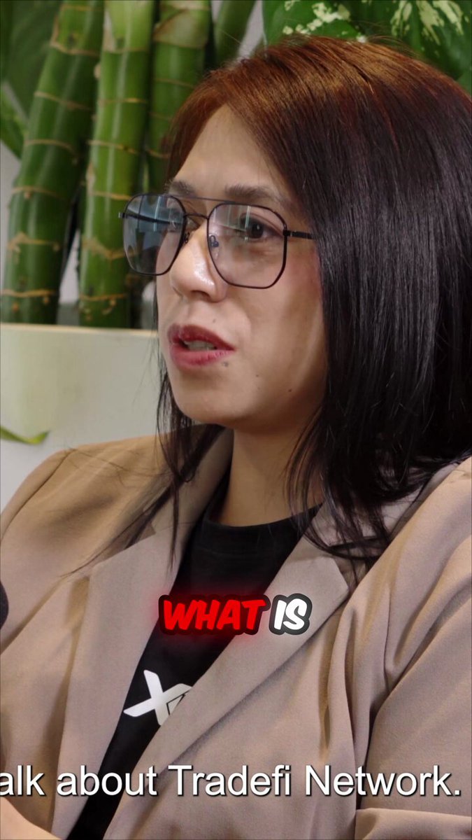 The image shows a woman with dark hair wearing glasses and sitting down in front of a green plant, holding an item. She appears to be engaged in conversation, as indicated by her facial expression. The background is blurred, focusing attention on the woman and the object she holds. The focus of the image is the woman, while the details related to the post are in the background, with a headline reading "Talk about TradeFi Network: #RWA #DeFi #Crypto".
