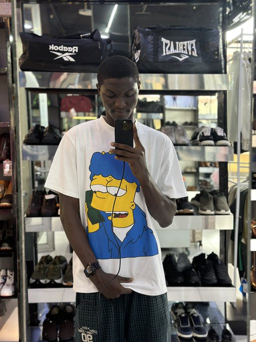 A young man takes a mirror selfie in a sneaker store wearing a white Bart Simpson t-shirt, with shelves of Reebok and Everlast shoes visible behind him.