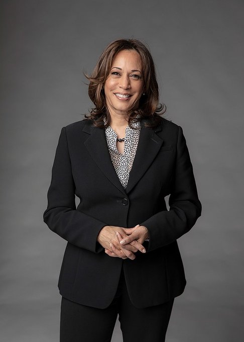 I can see a woman with shoulder-length brown hair, wearing a black blazer over a patterned blouse, smiling warmly with her hands clasped in front of her against a gray background. The image appears to be a professional portrait consistent with the post about Kamala Harris, who is pictured here in formal attire looking poised and confident—a fitting visual for the news about her potential 2028 presidential run and her new Pilates routine.