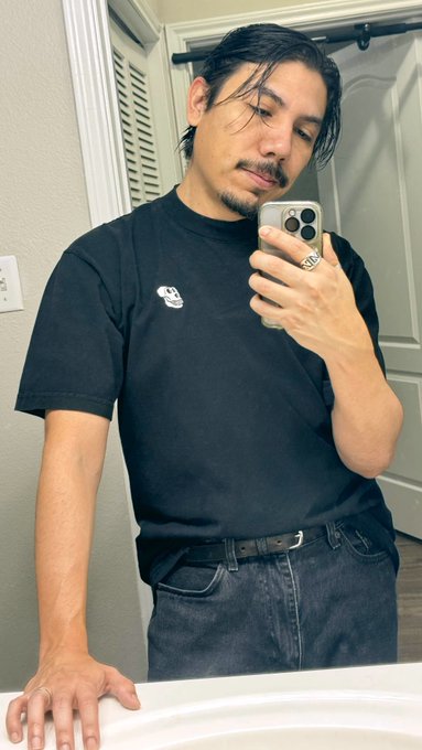A mirror selfie of a man with long dark hair wearing a black t-shirt, black jeans with a belt, and a silver ring, holding a smartphone in a bathroom.