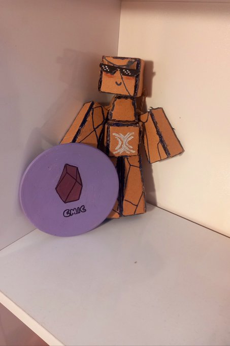 A small orange cardboard robot figurine wearing drawn-on sunglasses, holding a purple circular disc with a gem design and 'GMMTV' text, sitting in a corner.