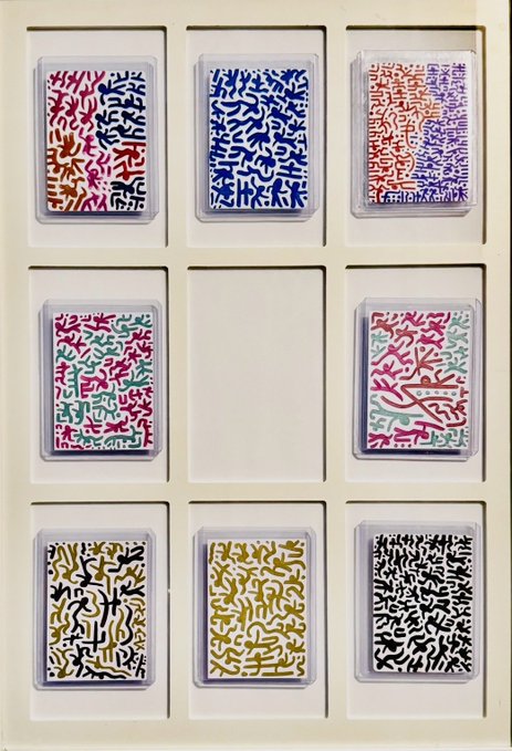 A cream-colored gridded frame displaying eight colorful abstract artworks with squiggly, organic patterns in various color combinations (pink/black, blue/white, purple, red, pink/green, yellow/black, gold, black/white), with one empty slot in the middle.
