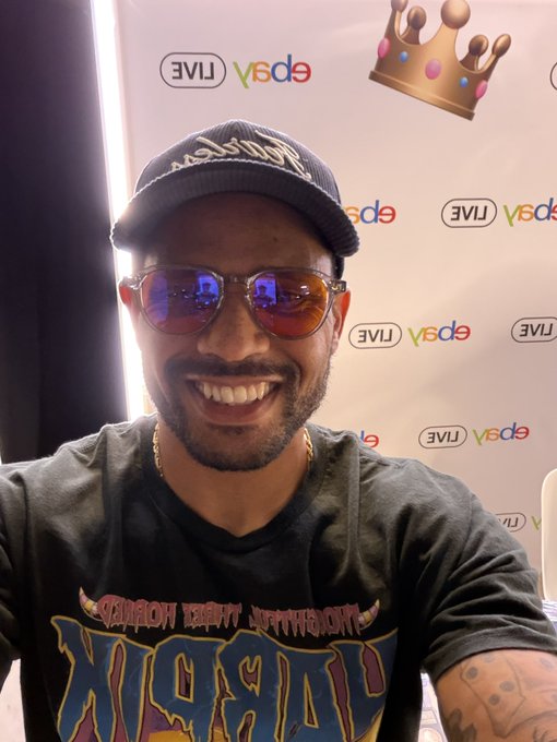 A smiling man wearing a black 'Lakers' cap, reflective sunglasses, and a graphic t-shirt poses at what appears to be an ebay LIVE event backdrop, with a crown emoji overlaid.